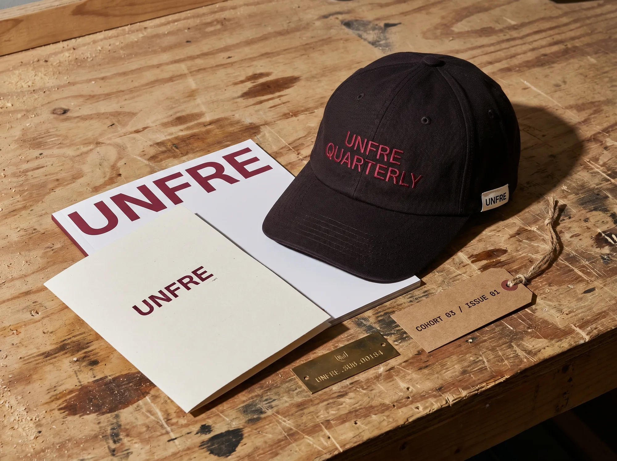 Unfre Quarterly Cap (Repair Culture & Anti-Disposability Editorial) — A subscriber-issue cap — heavy deep-charcoal canvas with UNFRE QUARTERLY embroidered in claret across the front panel.